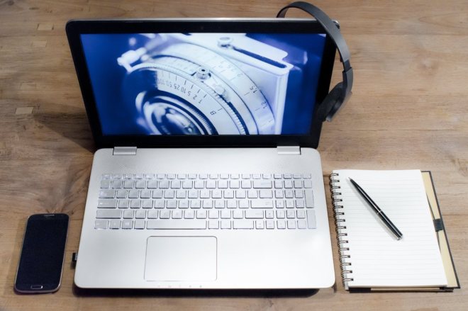 En smartphone, en laptop och ett anteckningsblock med en penna på ligger på ett bord. Laptopen har skärmen uppfälld och på skärmens högerkant hänger ett par hörlurar.